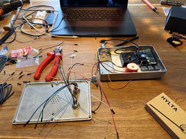 A busy workbench: the lid with buttons and loose wires, red pliers, and the open box with the speaker and Pi inside