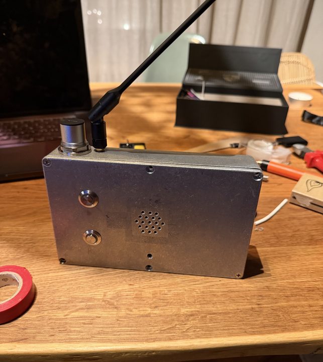 The finished box standing on its edge on a table: two stainless buttons on the left, a speaker grille in the middle, a USB-C socket and a black antenna on top