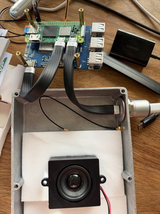 The Pi Zero on the modem board with ribbon USB cables, beside the open box with the speaker on a foam block and the antenna pigtail