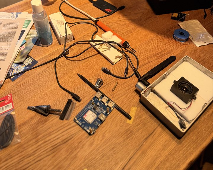 Parts spread on a wooden table: the LTE modem board, a step drill, a header strip, a soldering iron, and the open aluminium box with the speaker inside